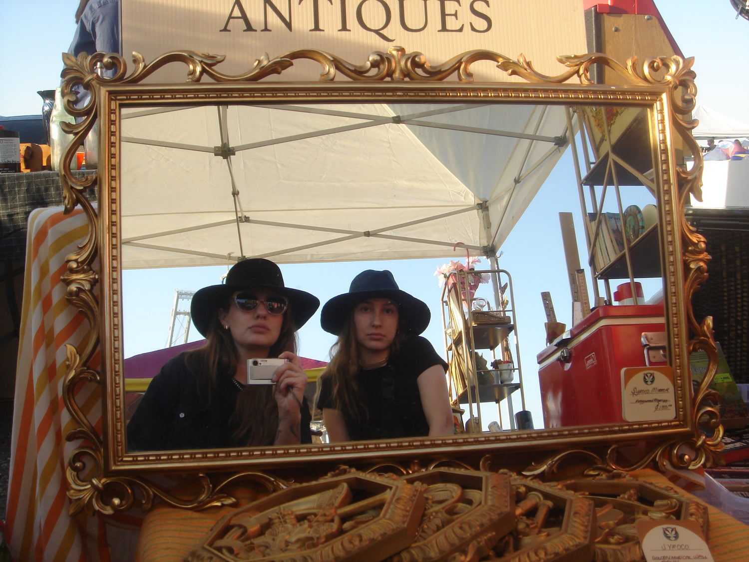 A large ornate gold-framed mirror on a vintage-style display stand at an antique store. The mirror reflects two people wearing black wide-brim hats, with a white canopy tent and outdoor market elements visible in the background.